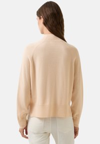 Vrouw met lang bruin haar, gekleed in een beige gebreide trui en witte broeken, gezien vanachter tegen een effen achtergrond.