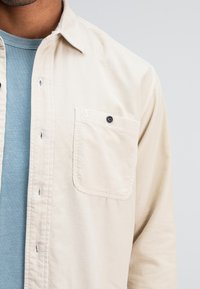 Beige corduroy shirt met een zak voorzien van een zwarte knoop en een geborduurd label. Gelaagd over een lichtblauw geribbeld shirt.