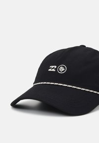 Gorra de béisbol negra con logotipo y insignia bordados, que presenta una banda decorativa de color blanco cosida alrededor del borde y tela texturizada.