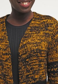 Virkat cardigan i orange och svarta nyanser, texturerat mönster, öppen front, med lös passform och randig svart topp under.