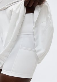 Isik kannab valgeid kõrge vöökohaga rattapükse küljetaskutega ning tõmbab valget soonikkoes rõivast vöökohtade kohalt üles.