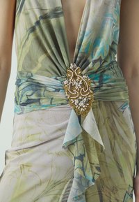 Mångfärgad klänningsomslag i genomskinligt tyg med blommönster, med en guldfärgad dekorativ brosch med pärlor och paljetter vid midjan.