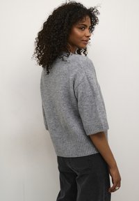 Pilkas megztinis trumpomis rankovėmis ir ribiniu apačios raštu. Minkšta tekstūra ir laisvas siluetas, derinamas su tamsiais džinsais, priešais vientisą foną.