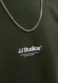 T-shirt en coton vert foncé portant un logo imprimé blanc "JJ Studios" et un collier en chaîne métallique argenté drapé autour de l'encolure.