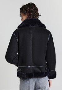 Person mit kurzen, gewellten Haaren, der eine schwarze Lammfelljacke mit Fellkragen und -manschetten trägt und vor einem schlichten, hellen Hintergrund steht.