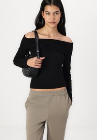 Černý svetr bez ramínek s žebrovaným vzorem a dlouhými rukávy, spárovaný se světle béžovými kalhotami s elastickým pasem. Modelka nese malou kulatou tašku se cvočky.