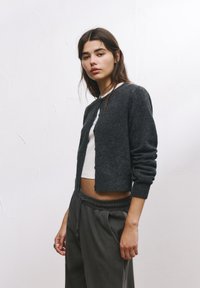Cardigan din lână de culoare gri închis, cu un design scurtat, guler rotund și deschidere cu butoane, purtat peste o cămașă albă și asortat cu pantaloni de trening gri.