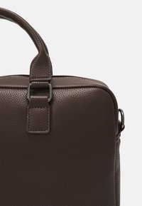 Sac en cuir marron avec une surface texturée, doté d'une poignée supérieure et d'une bandoulière amovible. Comprend un détail en boucle métallique sur le rabat.