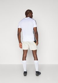 Hvid atletisk skjorte med sorte sidepaneler, parret med lysebeige shorts. Modellen bærer sorte fodboldstøvler og hvide sokker, hvilket viser et sporty design.
