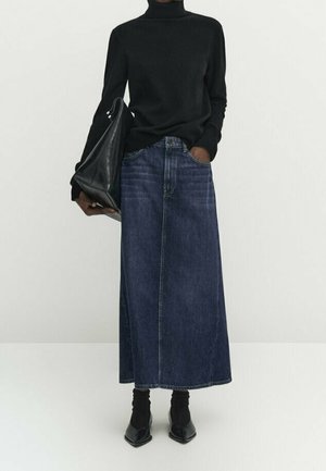 Personne portant un col roulé noir, une longue jupe en jean foncé, des chaussures noires pointues, tenant un grand sac à main en cuir noir, debout devant un fond uni.