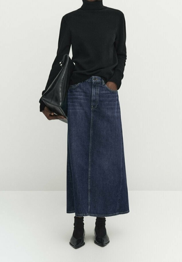 Personne portant un col roulé noir, une longue jupe en jean foncé, des chaussures noires pointues, tenant un grand sac à main en cuir noir, debout devant un fond uni.