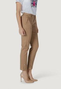 Pantalon beige à coupe ajustée, avec poches latérales, plis à l'avant et poignets boutonnés, associé à des talons hauts couleur nude et un t-shirt blanc avec imprimé.