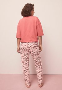 Světlé růžové bavlněné tričko s krátkými rukávy, kombinované s bílými kalhotami s červeným srdíčkovým vzorem, pohled zezadu.