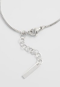 Zilveren metalen ketting met een flexibele schakels ontwerp, een karabijnsluiting en een verstelbare verlenging met een rechthoekig label waarin "STEE" is gegraveerd.