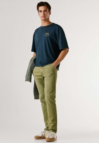 Tmavomodré bavlnené tričko so zlatým logom, olivovo zelené nohavice a tenisky s vzorom. Ľahký sveter prehodený cez rameno, úzky strih.