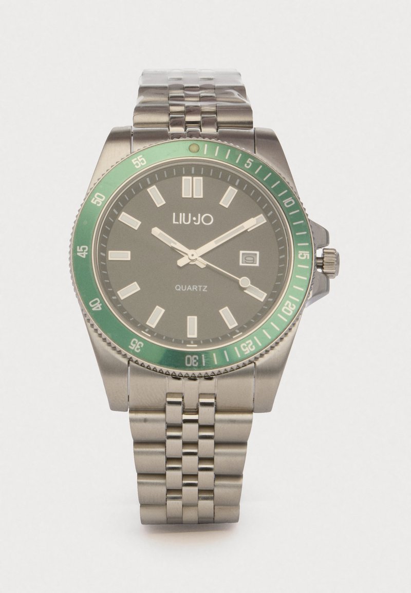 Kellotaulu, jossa on vihreä kehys, harmaa tausta ja hopeiset yksityiskohdat. Varustettu kvartsikoneistolla ja päivämääränäytöllä kello 3 kohdalla.