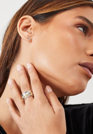 Close-up of a woman touching her neck, wearing a small heart-shaped earring and three mixed-metal textured rings on her finger.