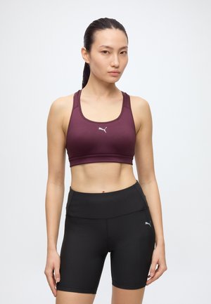 Femme aux cheveux foncés en queue de cheval, portant un soutien-gorge de sport bordeaux et un short de cyclisme noir taille haute sur un fond blanc uni.