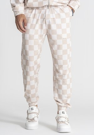 Osoba nosiaca béžové a biele kárované teplákové nohavice s elastickými manžetami a biele tenisky s logom na jazyku, stojaci s rukami v vreckách.