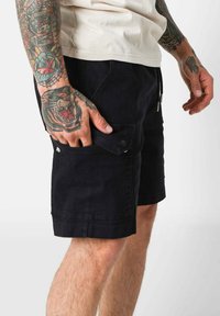 Pantaloni corti neri realizzati in tessuto resistente, con una tasca laterale con chiusura a scatto e vita con coulisse. Texture liscia, design alla lunghezza del ginocchio.