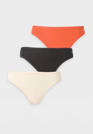 Mujer con cabello largo y oscuro, que lleva un bikini naranja, de pie frente a un fondo claro liso, mirando hacia adelante con una expresión neutral.