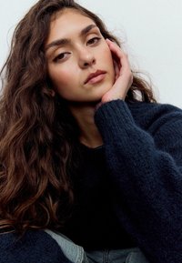 Primo piano di una persona con lunghi capelli scuri e mossi, che indossa un maglione scuro dalla trama marcata e un capo in denim chiaro. Sfondo morbido e neutro.