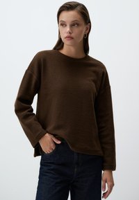 Felpa marrone in pile con colletto rotondo e maniche lunghe, caratterizzata da una vestibilità comoda e spacchi laterali. Abbinata a jeans in denim scuro.