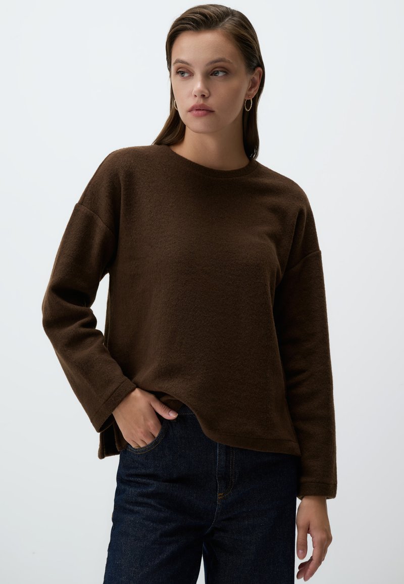 Felpa marrone in pile con colletto rotondo e maniche lunghe, caratterizzata da una vestibilità comoda e spacchi laterali. Abbinata a jeans in denim scuro.