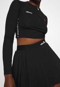 Czarna długa bluzka z rękawami do łokci, croptop z zamkiem błyskawicznym, ozdobiona naszywkami z logo. Połączona z czarną plisowaną spódnicą, obie wykonane z gładkiej, elastycznej tkaniny.