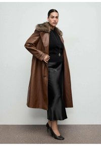 Manteau en cuir marron avec un col en fourrure, assorti à un haut noir et une jupe noire en satin. Des talons noirs complètent la tenue.
