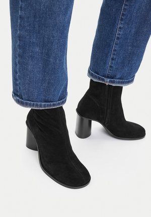 Black suede ankle boots with block heels worn with cuffed blue jeans on a white background.