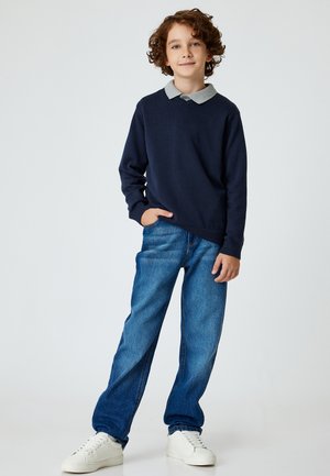 Marineblauer Pullover mit grauem Kragenakzent, kombiniert mit blauen Jeans und weißen Sneakers. Der Pullover hat einen lockeren Schnitt und einen lässigen Stil.