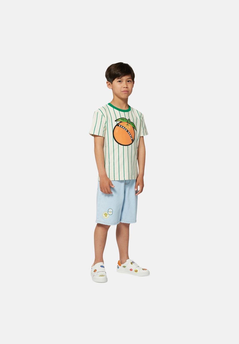 T-shirt di cotone con strisce verdi e grafiche arancioni, abbinato a pantaloni shorts in denim blu chiaro e sneakers bianche con vari patch.