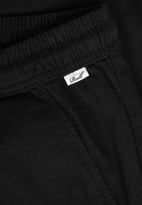 Pantalones con cintura elástica negra y una pequeña etiqueta blanca con la palabra "Reell" cosida cerca de la costura del bolsillo.