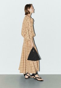 Beige geruite jurk met losse mouwen, vastgebonden in de taille, gecombineerd met zwarte sandalen en een driehoekige zwarte tas. Gladde textuur, vloeiend ontwerp.