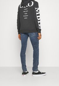 Tmavě šedá mikina s kapucí a bílým textem uvádějícím města, v kombinaci s modrými džíny a černými teniskami s bílou podrážkou.