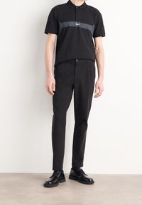 Černé polo tričko s šedým horizontálním pruhem, v kombinaci s černými zeštíhlenými kalhotami a lesklými černými botami; minimální design s knoflíkovým límcem.