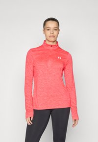 Röd långärmad träningsblus med halvdragkedja, tillverkad av strukturerat tyg, med en liten logotyp på bröstet. Bärs med svarta tights.