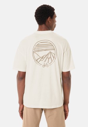 Uomo visto di spalle che indossa una maglietta color avorio con una grafica sul retro che raffigura una montagna, una bicicletta e il sole in marrone chiaro.