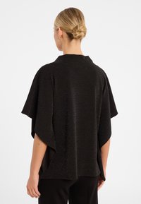 Schwarzer, offener Kimono mit kurzen Ärmeln, aus strukturiertem Stoff mit goldenen Sprenkeln. Der abgerundete Saum und die seitlichen Schlitze verbessern die Beweglichkeit.