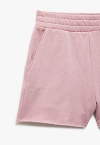 Pantalones cortos de color rosa claro con una textura suave, cinturilla elástica con costuras verticales y bolsillos laterales. El dobladillo desmechado añade un detalle casual.