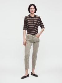 Femme aux cheveux courts et foncés portant un haut rayé marron, un pantalon beige et des ballerines noires, debout avec une main dans la poche sur un fond uni.