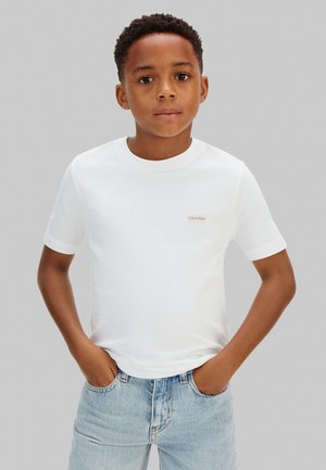 Băiat în picioare cu mâinile în buzunare, purtând un tricou alb Calvin Klein și blugi deschis la culoare, în fața unui fundal gri.