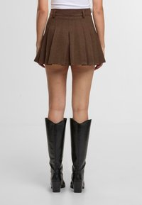 Hnědá plisovaná mini sukně vyrobená z texturované látky, zobrazená ze zadní strany, v kombinaci s černými koženými botami po kolena.