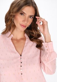 Blusa rosa con una padding, dettagli in argento e oro, colletto arruffato e maniche a sbuffo. I dettagli con bottoni arricchiscono il design. Tessuto morbido e leggero.