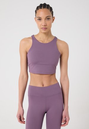 Femme aux cheveux tressés portant un crop top violet sans manches et un legging taille haute, debout devant un fond blanc uni.