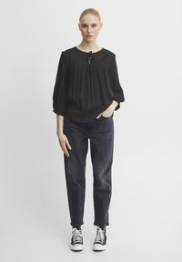 Blusa nera leggera con scollatura rotonda, dettaglio con lacci e vestibilità ampia. Abbinata a jeans scuri e sneakers alte.