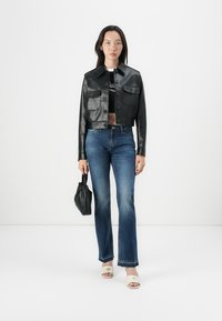 Svart lær cropped jakke med to frontlommer, kombinert med denimjeans. Modellen holder en liten svart veske og har på seg hvite åpne hæler.