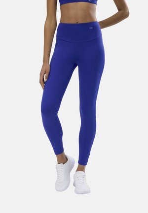 Mujer con leggings de cintura alta azules y sujetador deportivo a juego, de pie con zapatillas deportivas blancas sobre un fondo liso.