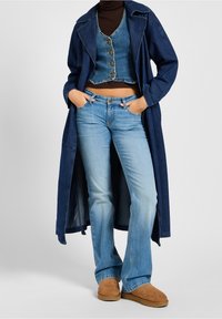 Trench coat de denim en azul oscuro, superpuesto a un chaleco de denim en azul claro. Jeans acampanados en azul claro y botines de ante marrón.